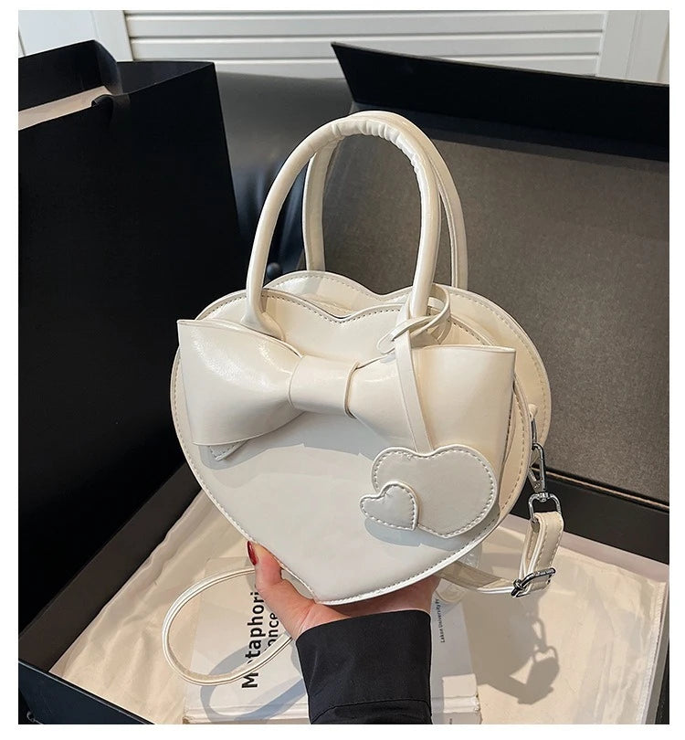 carauana-lovely-shoulder-bag-heart White heart-shaped handbag with a bow detail held by a person.